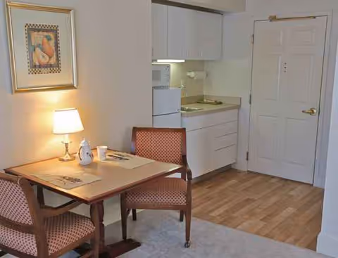 Small dining area with table, two upholstered chairs, lamp, artwork, and a kitchenette in Cove's Edge.