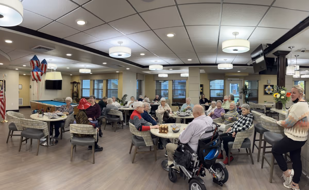A group of elderly people sitting around tables in a spacious, well-lit common area with modern decor. Some are engaged in conversation, and one person is in a wheelchair. There are American flag balloons and a pool table in the background. A woman stands on the right side of the image, smiling.