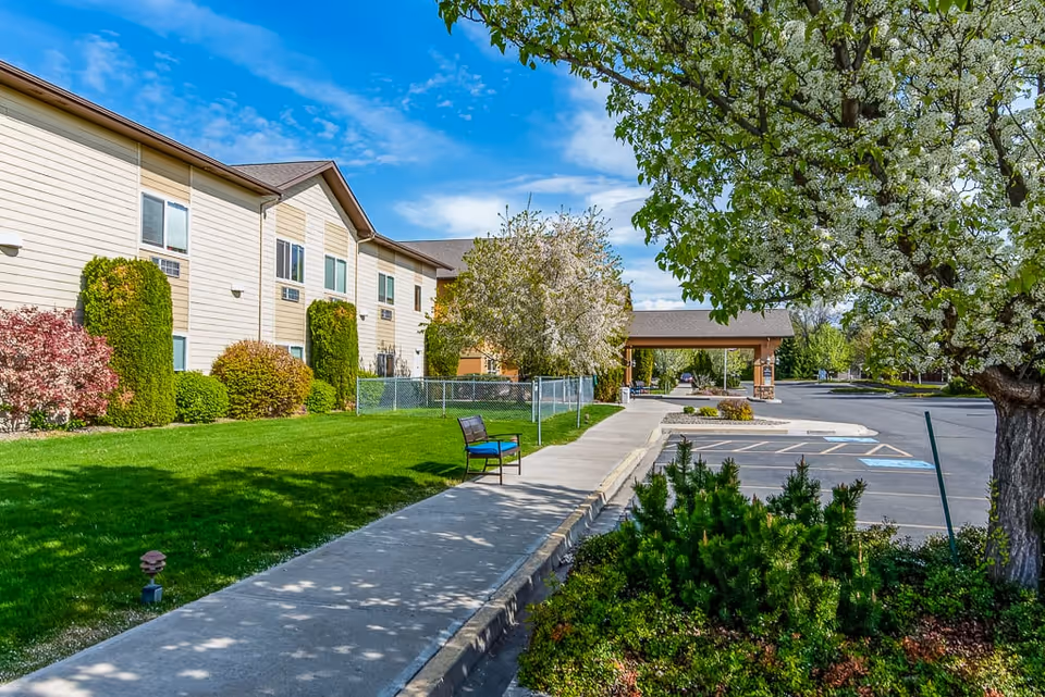 Exterior view of Ellensburg Senior Living facility showing a two-story beige building with multiple windows, a well-maintained green lawn, various bushes and trees in bloom, a sidewalk with a bench, and a parking lot with marked spaces under a blue sky with scattered clouds.