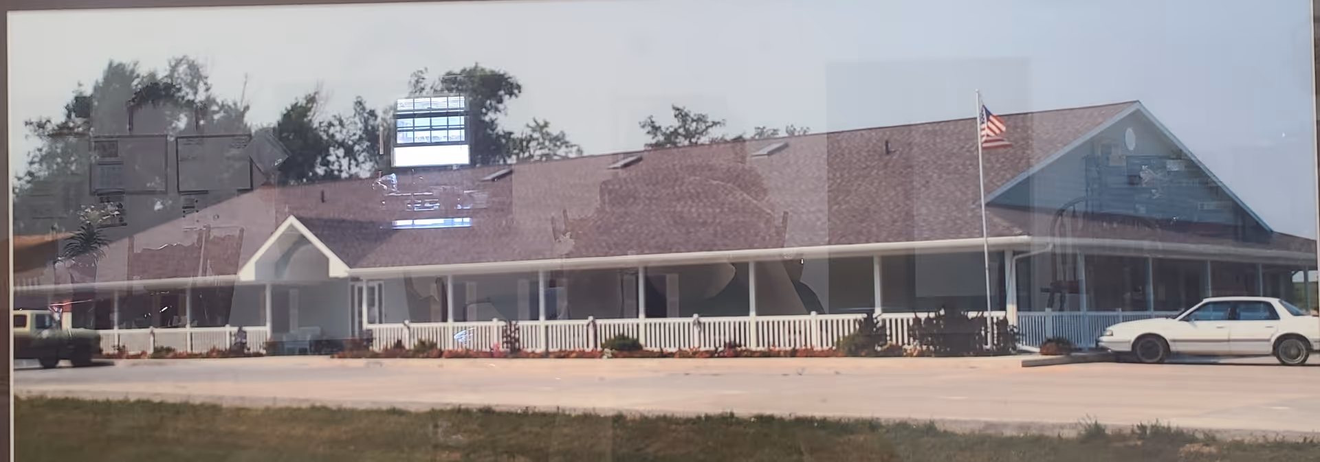 Exterior view of a single-story assisted living facility with a long porch, white railing, and an American flag on a flagpole in front. There are two cars parked near the building and some trees in the background.