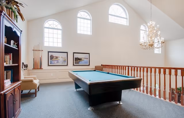 A bright recreational room with a pool table in the center, carpeted floor, and a wooden railing overlooking a lower level. The room features three large arched windows letting in natural light, a chandelier hanging from the ceiling, framed landscape paintings on the wall, a wooden bookshelf with books and decor, and a beige armchair.