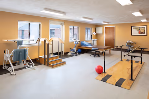A bright rehabilitation room with parallel bars, therapy stairs, exercise equipment, a treatment table and exercise balls.