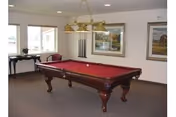 A recreation room with a red-felt pool table under hanging lights, framed artwork on the walls, windows, and a small table along the wall.
