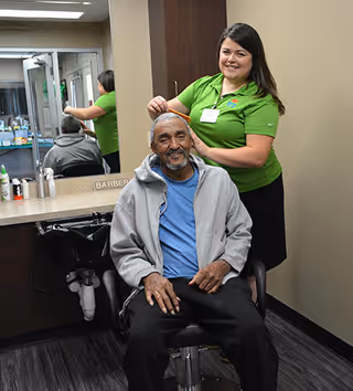 A smiling elderly man sits in a barber chair wearing a gray hoodie and blue shirt while a woman in a green shirt stands behind him, combing his hair in a salon area with a mirror and sink visible.