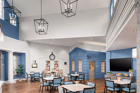 Bright, high-ceiling communal dining area with multiple tables and chairs, blue accent walls and decorative small house-like structures.