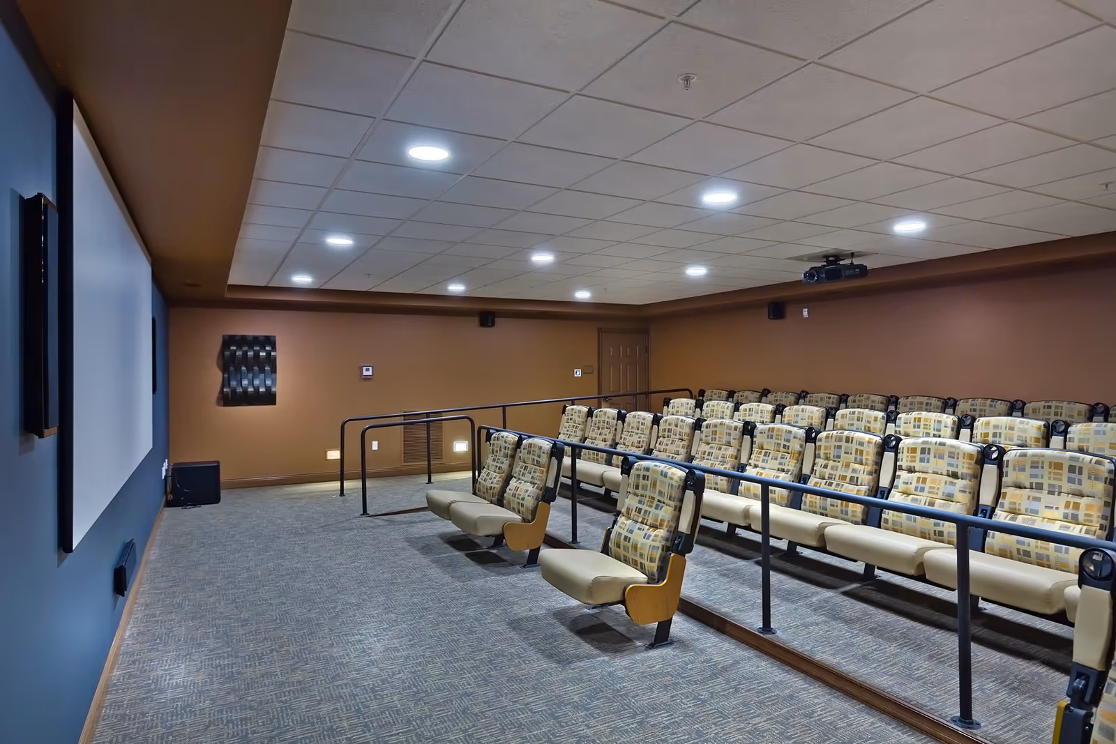 Interior view of a small theater room with rows of cushioned seats featuring patterned upholstery. The room has a large projection screen on the left wall, recessed ceiling lights, and brown walls. There are handrails along the aisles and a projector mounted on the ceiling.