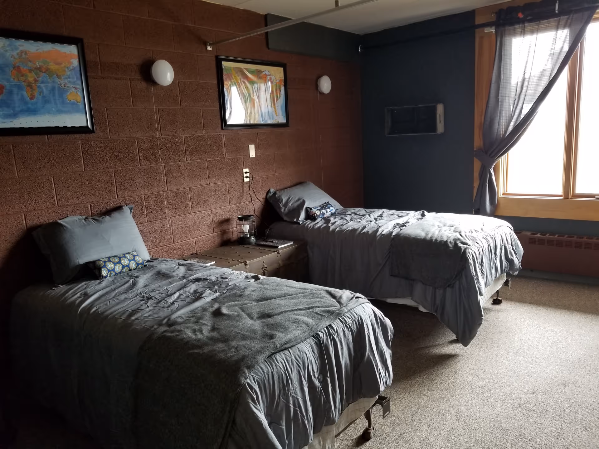 A bedroom with two single beds covered in gray bedding. Between the beds is a small trunk used as a nightstand with a lantern and a book on it. The walls are painted dark brown and dark blue, with two framed maps hanging on the brown wall. A large window with sheer gray curtains lets in natural light.