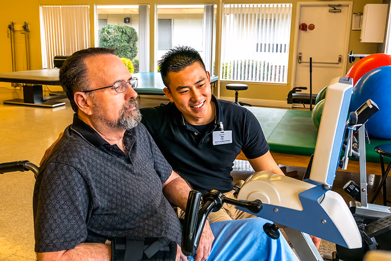 A healthcare professional assists an elderly man in a wheelchair with a physical therapy exercise using a specialized machine in a bright therapy room with large windows and exercise equipment.
