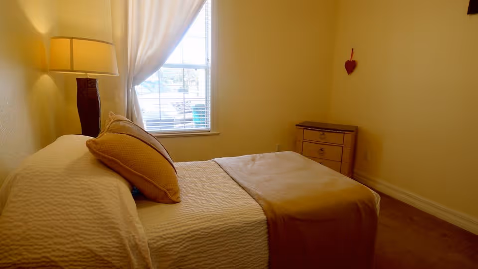 A cozy bedroom with a neatly made bed featuring white bedding and a beige throw blanket. There are two beige pillows on the bed. A wooden nightstand with a lamp is positioned next to the bed. A window with white curtains allows natural light to brighten the room. A small wooden dresser is placed against the far wall, which is decorated with a small red heart hanging.
