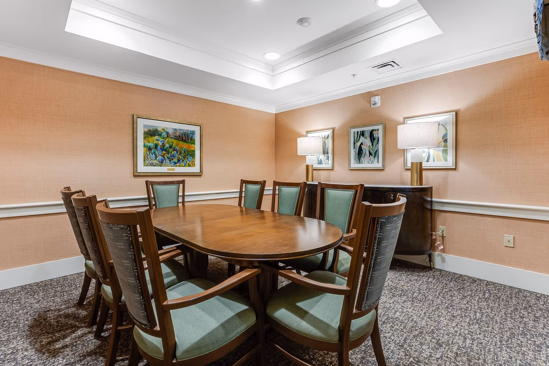 A small dining/meeting room with an oval wooden table surrounded by upholstered chairs, framed artwork, and table lamps against peach-colored walls.