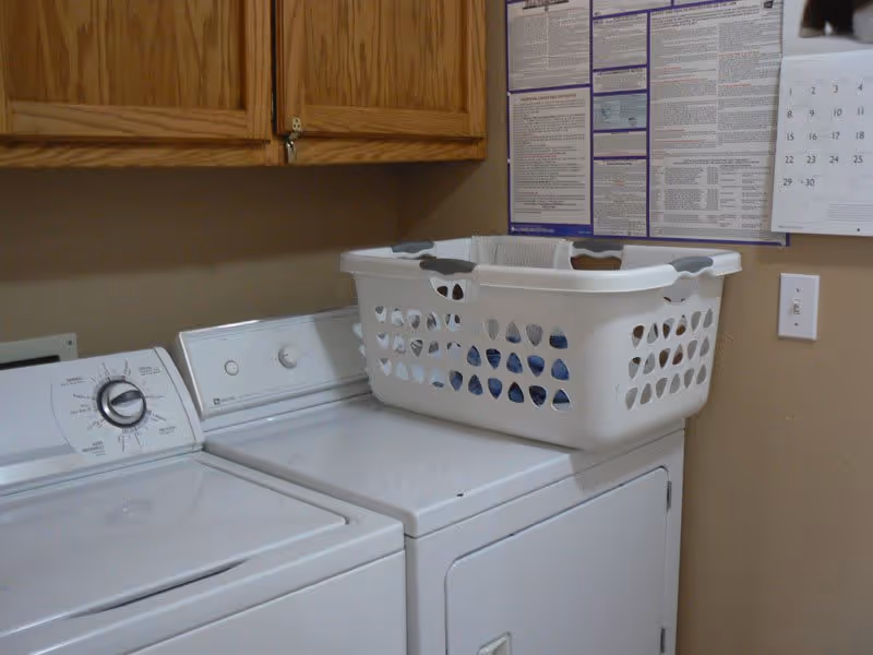 Laundry room with a white washing machine and dryer side by side. A white laundry basket is placed on top of the dryer. Above the machines, there are wooden cabinets mounted on the wall. A calendar and a large informational poster are visible on the beige wall to the right.