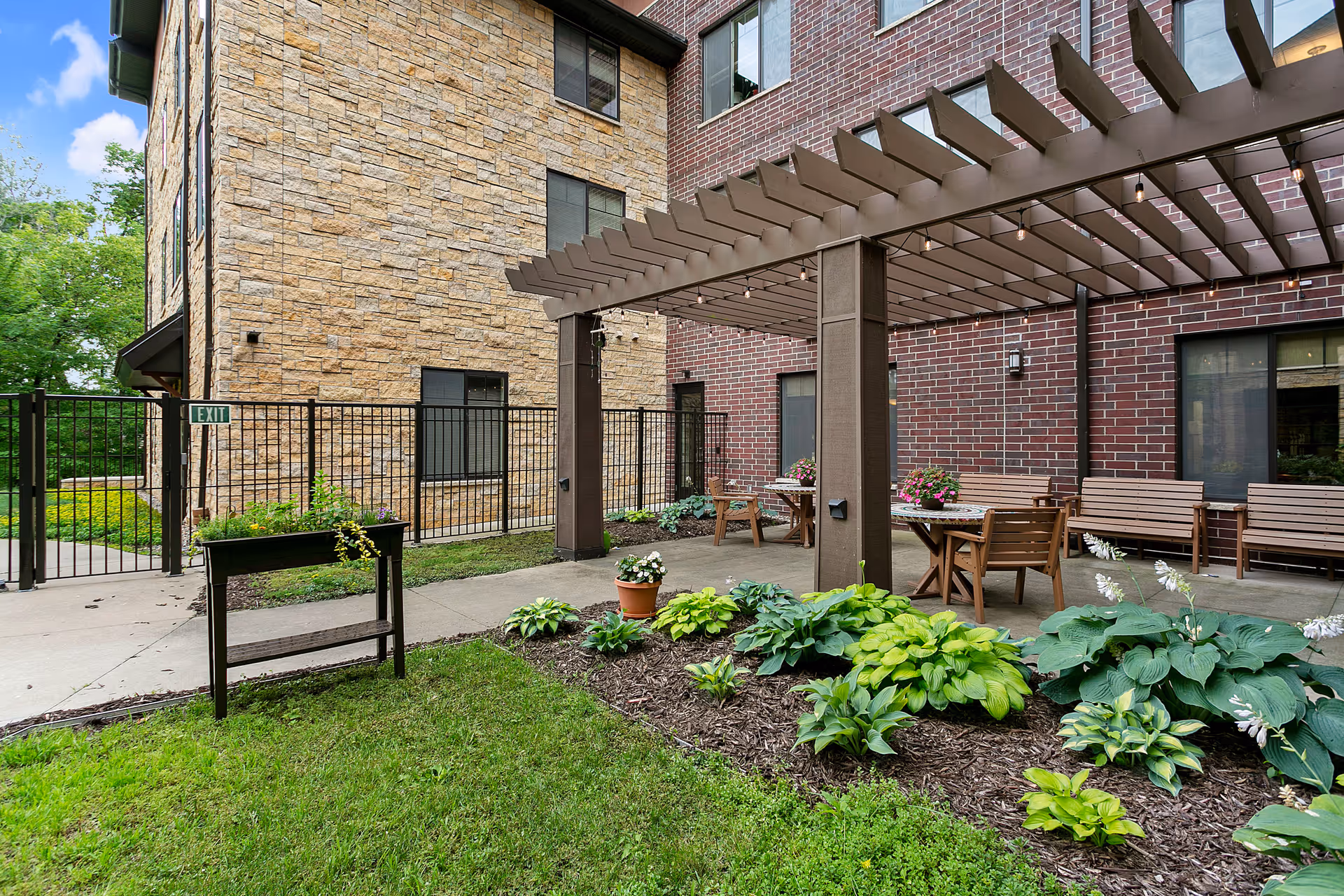 Outdoor courtyard with a pergola, patio tables and benches next to a brick and stone building and landscaped plant beds.