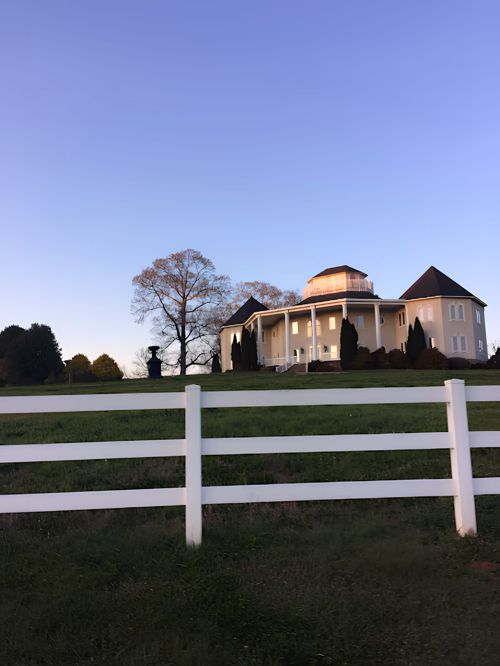 A large, elegant building with a circular section and multiple peaked roofs, surrounded by a white fence and green grass, under a clear blue sky during sunset or sunrise.