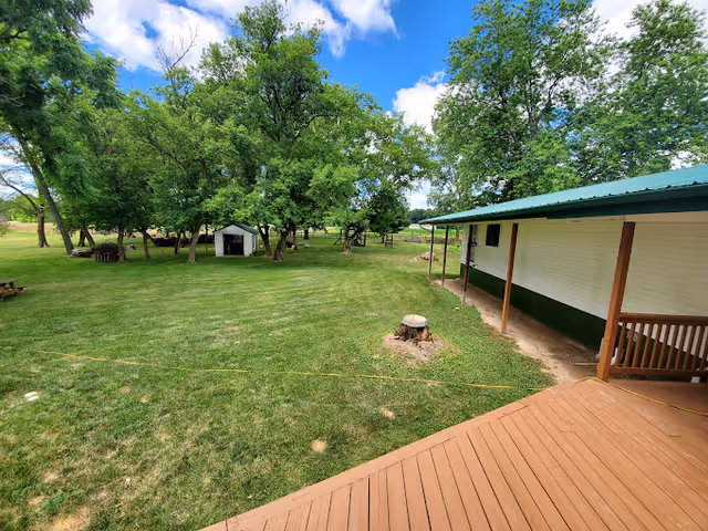 A spacious outdoor area with green grass, several trees, a small white shed, and a building with a green roof and a wooden deck in the foreground under a partly cloudy blue sky.