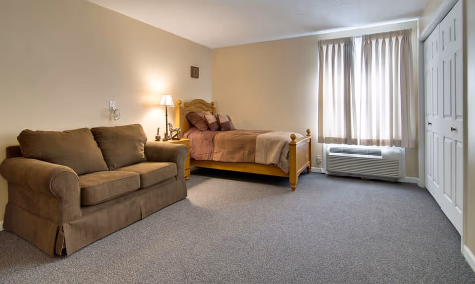 A senior living facility bedroom with a wooden bed dressed in brown bedding, a brown sofa, a nightstand with a lamp, beige walls, a window with light-colored curtains, and a carpeted floor.