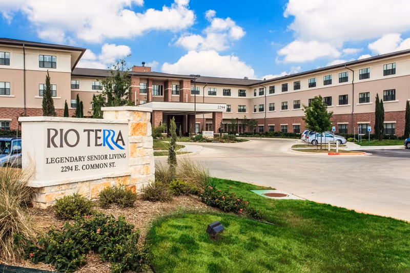 Exterior view of Rio Terra Legendary Senior Living facility showing a three-story building with beige and brick facade, a driveway, parked cars, landscaped greenery, and a stone sign with the facility name and address.