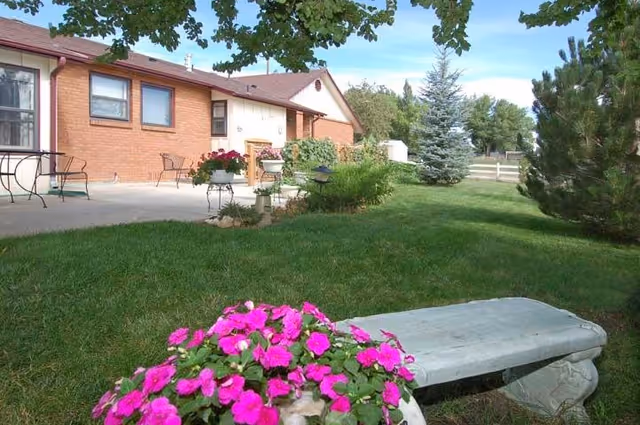 Small brick single-story building with a patio and lawn, a stone bench, and bright pink flowers in the foreground.
