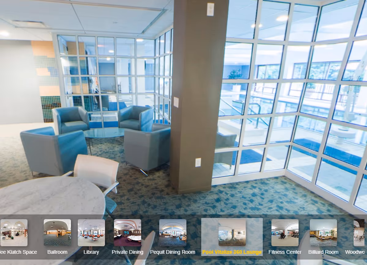 A bright lounge area with four blue armchairs arranged around a glass coffee table, adjacent to large windows overlooking an indoor swimming pool. There is a round table with chairs in the foreground and a carpeted floor with a floral pattern.