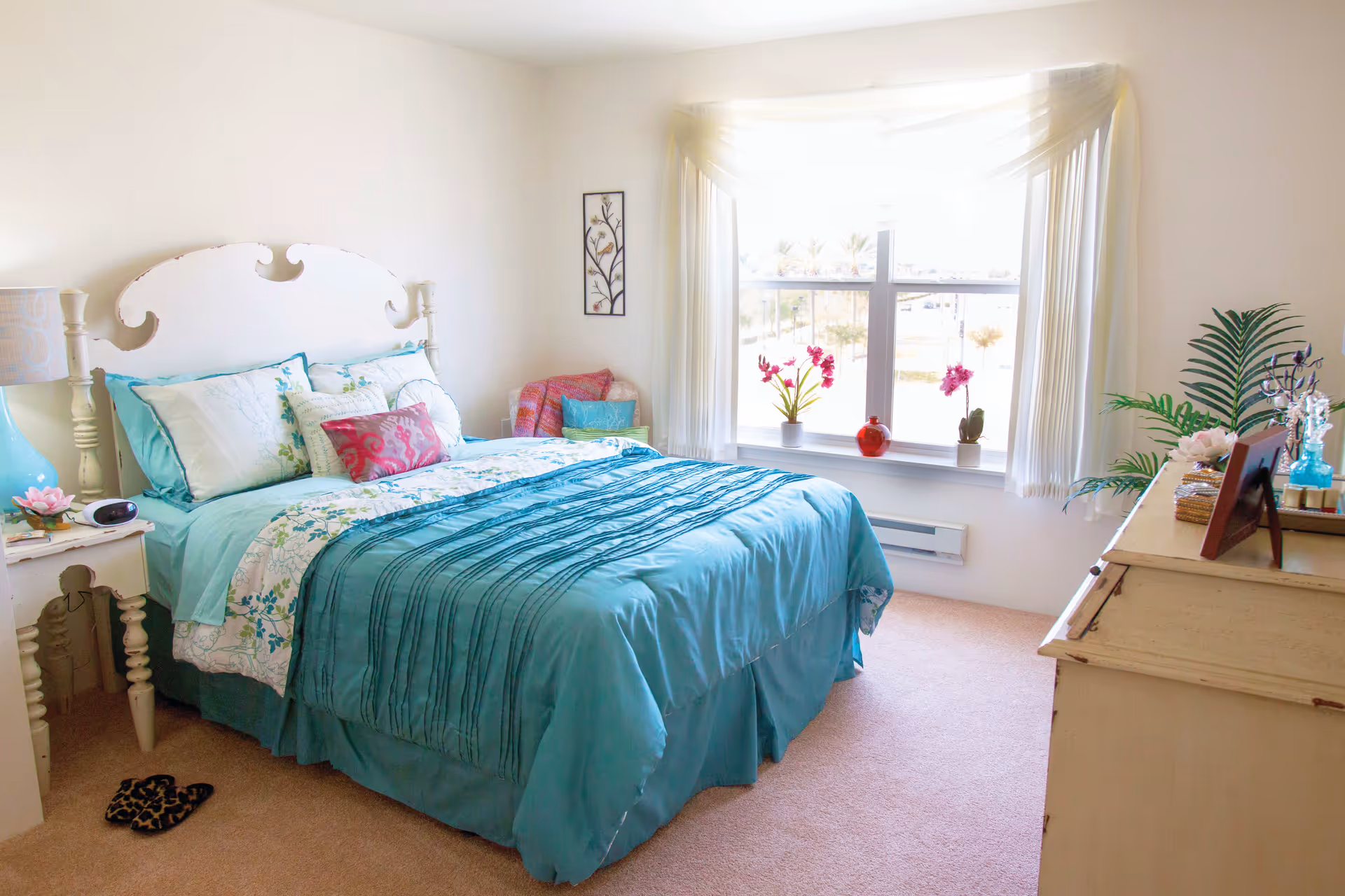 A bright and cozy bedroom with a large window letting in natural light. The bed has a white headboard and is made with teal and floral bedding, accompanied by multiple pillows. There is a white nightstand with a lamp and decorative items on the left side of the bed. A small chair with a pink cushion is placed near the window. On the right side, there is a dresser with plants, framed photos, and decorative pieces. The room has light-colored walls and carpeted flooring.