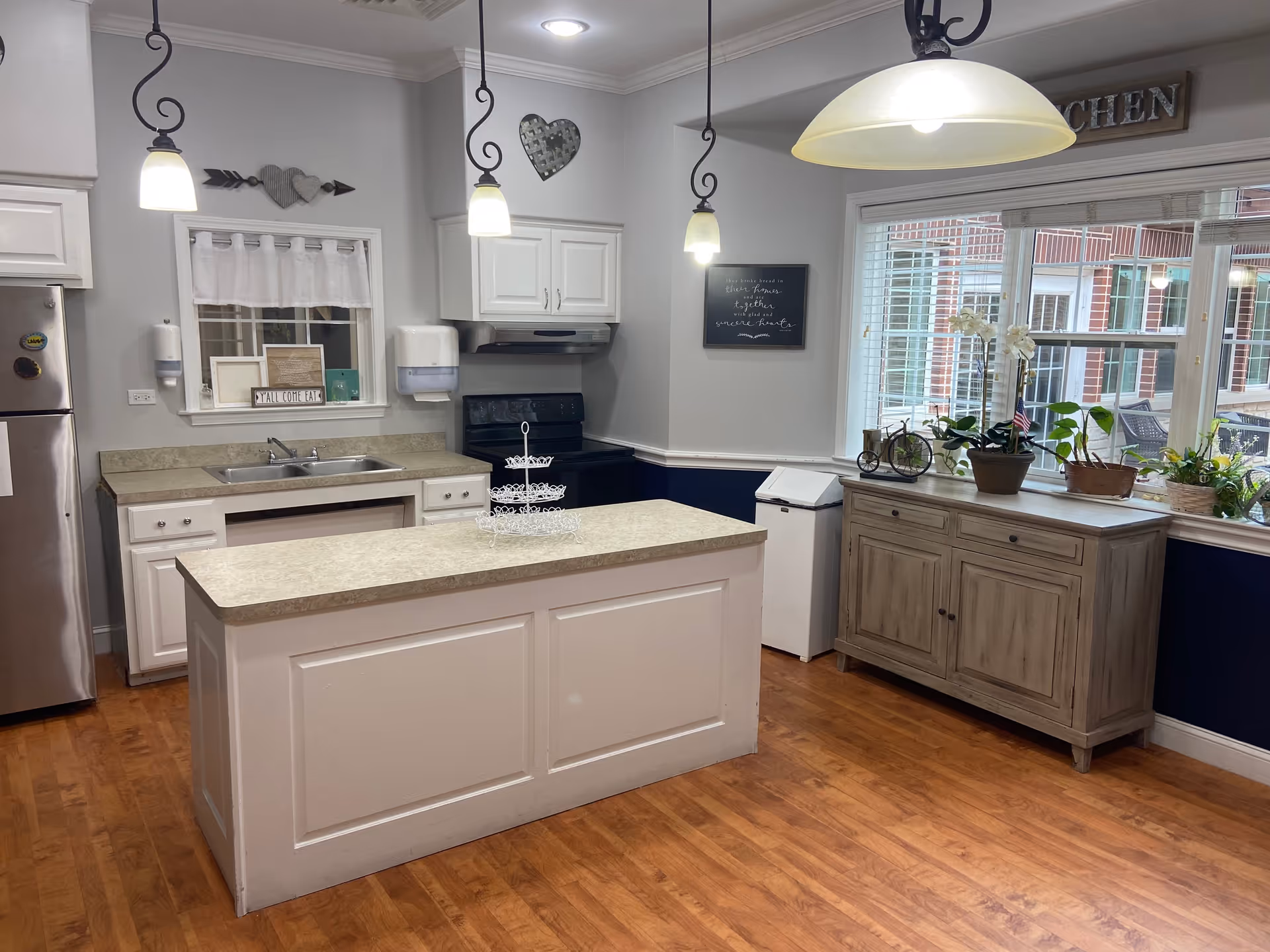 A bright kitchen with white cabinets, a central island with a beige countertop, stainless steel refrigerator, stove, and a window with white blinds. There are three pendant lights hanging from the ceiling and a wooden sideboard with potted plants and decorative items beneath a large window. The walls are painted white with a dark blue lower section and wooden flooring.