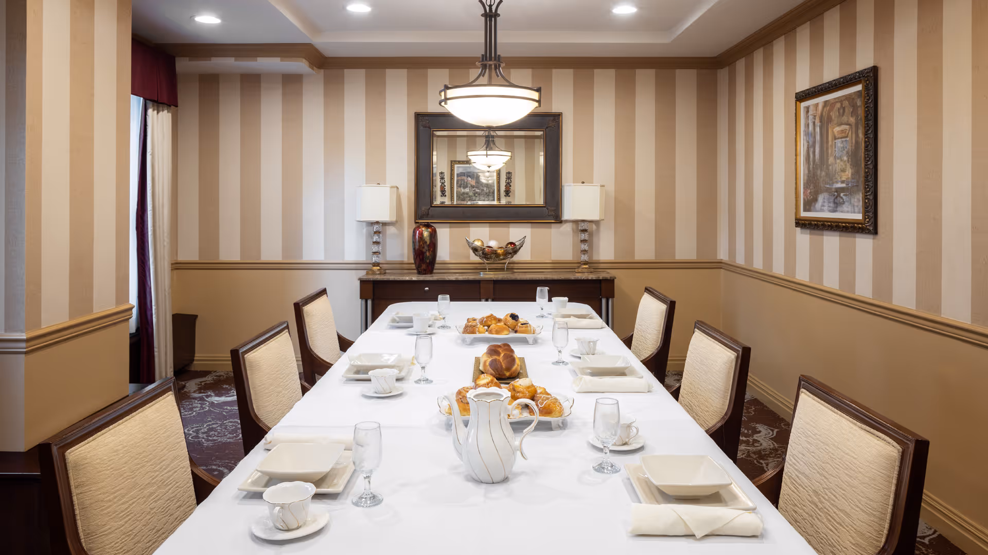 A formal dining room with a long table covered with a white tablecloth, set with plates, cups, glasses, and a centerpiece of pastries and a white pitcher. The room has striped wallpaper, a framed painting on the right wall, a large mirror on the back wall, two table lamps, and a chandelier hanging above the table.