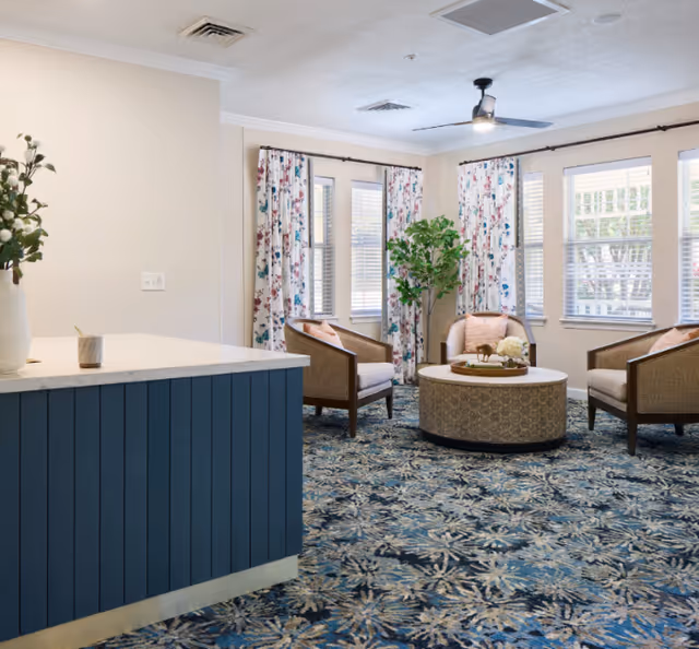 A cozy sitting area with four cushioned armchairs arranged around a round coffee table on a patterned carpet. The room has multiple windows with floral curtains, a ceiling fan, and a blue paneled counter with a vase of flowers on it.