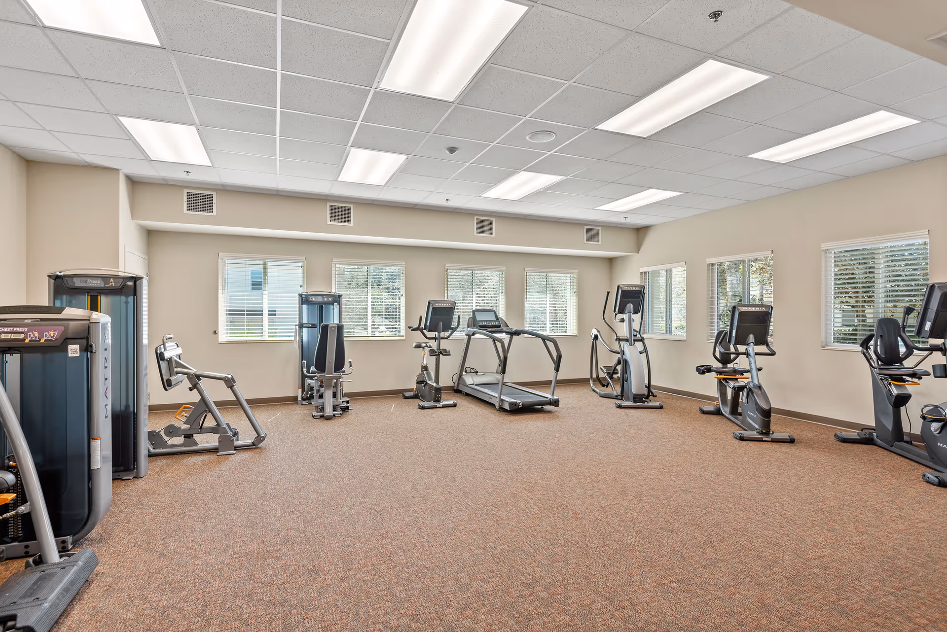 A spacious fitness room in a senior living facility with various exercise equipment including treadmills, stationary bikes, elliptical machines, and weight machines. The room has beige walls, a brown carpeted floor, and multiple windows with white blinds allowing natural light to enter.