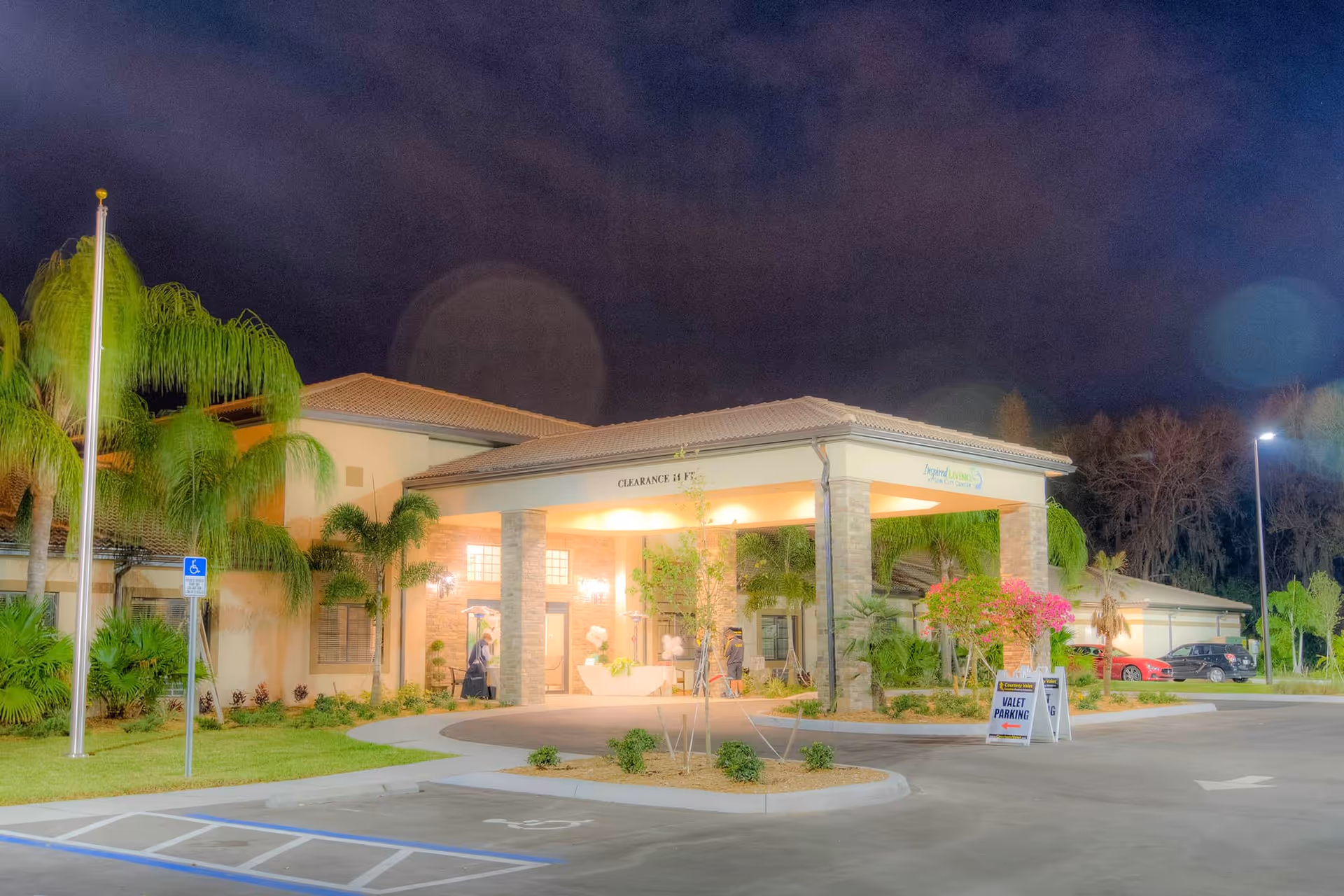 Night view of the entrance to Inspired Living Sun City Center, a senior living facility, with a covered drop-off area supported by stone pillars, palm trees, and a valet parking sign in the driveway.