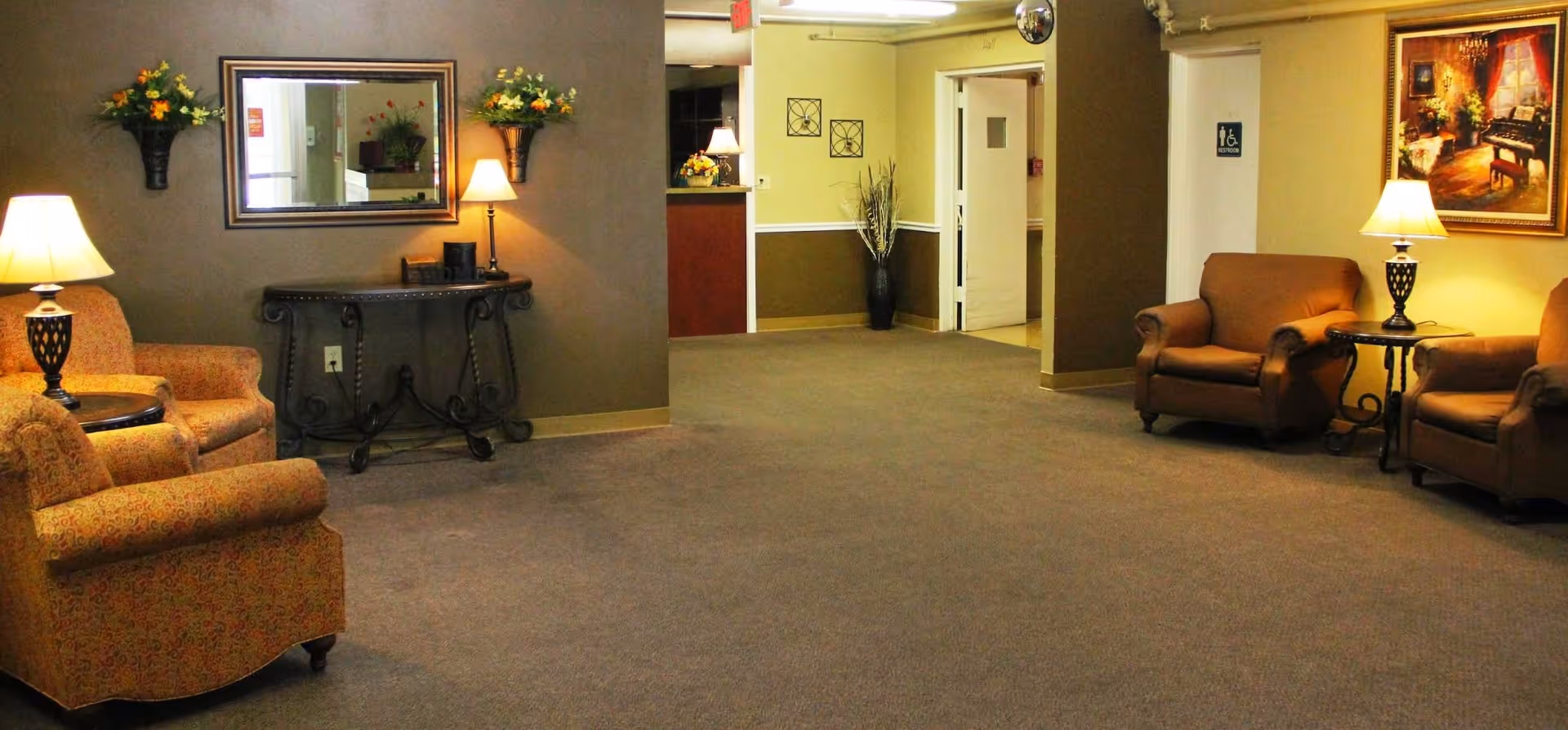 A cozy interior common area with carpeted floor, featuring four upholstered armchairs arranged around two side tables with lamps. The walls are decorated with a large mirror, floral wall sconces, and framed artwork. In the background, there is a hallway with a door marked with a handicap sign.