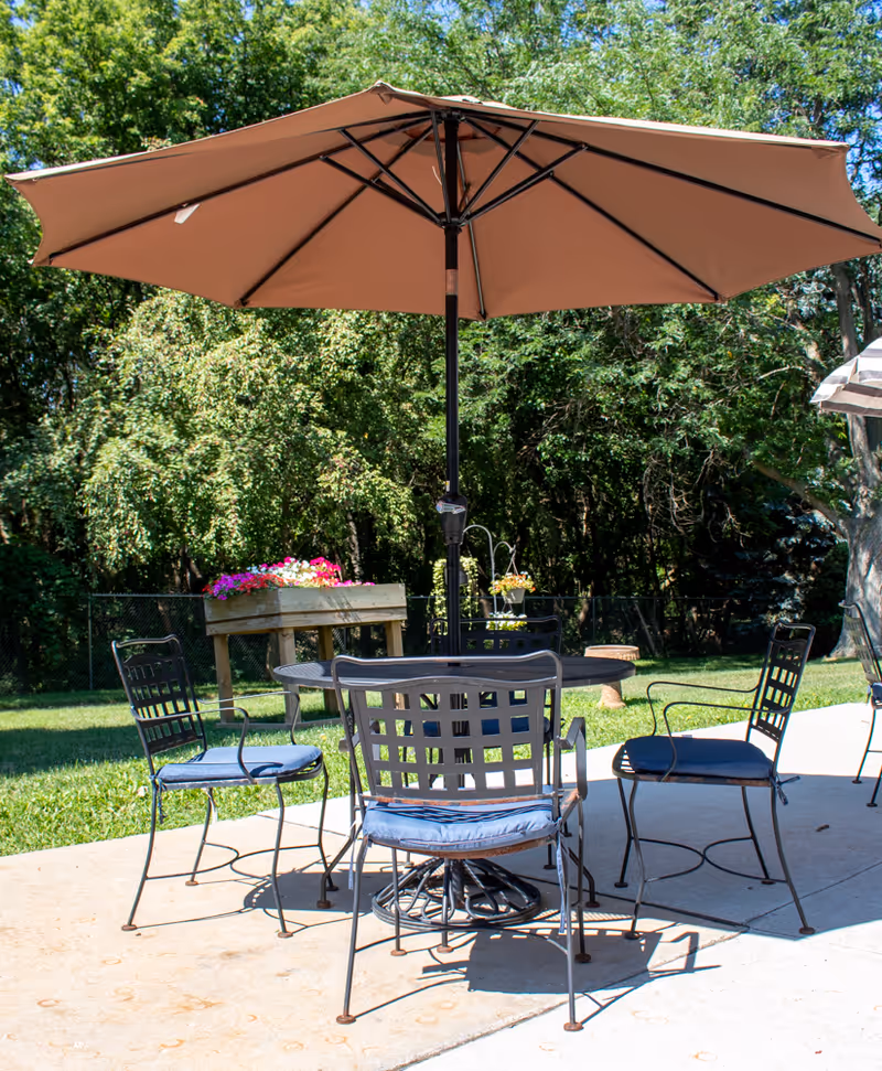 Outdoor patio set with a metal table and four cushioned chairs under a large brown umbrella on a concrete slab with trees and lawn in the background.