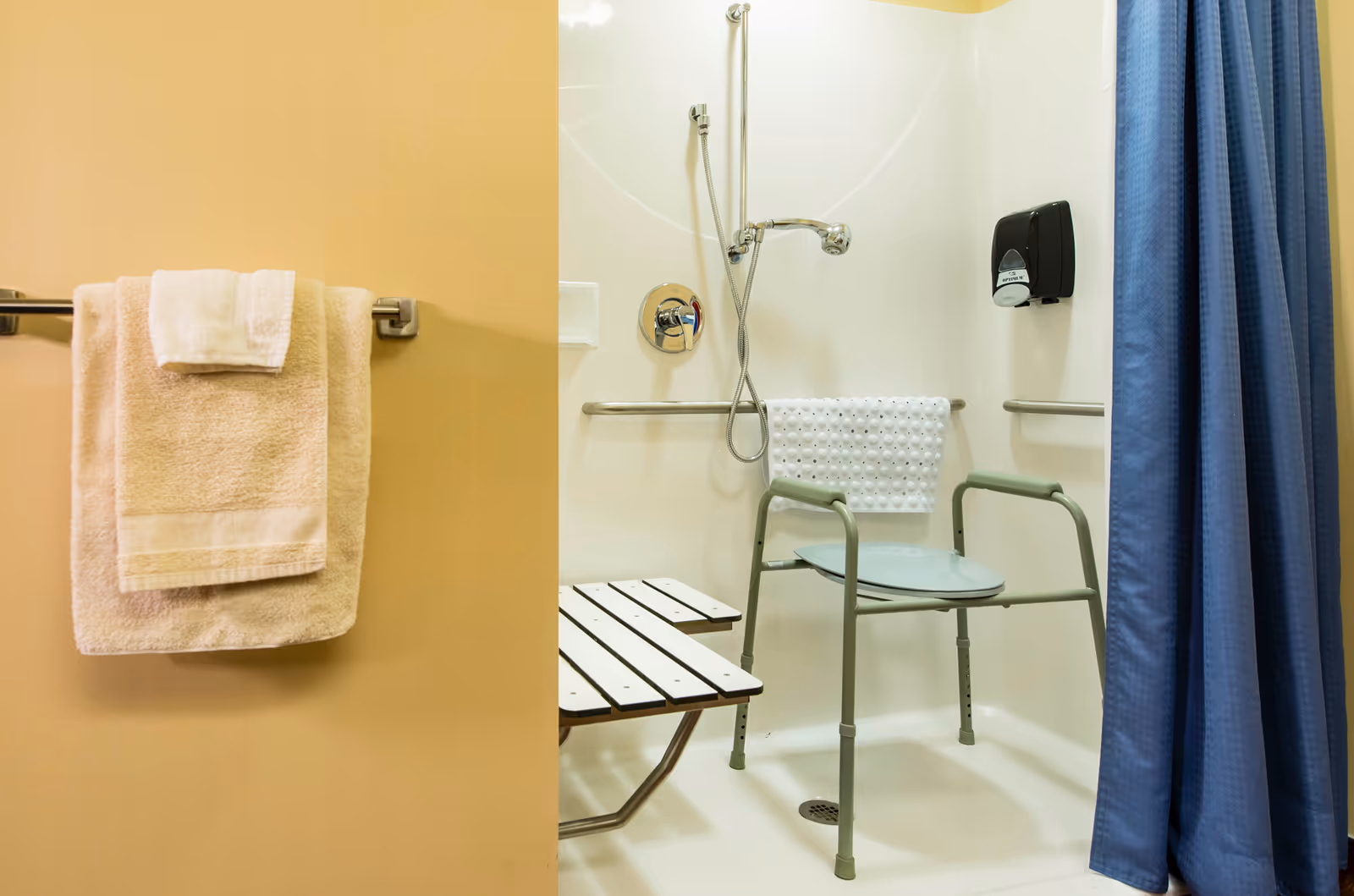 Accessible shower stall with a shower chair, grab bars, soap dispenser, blue curtain and towels on a nearby towel rack.
