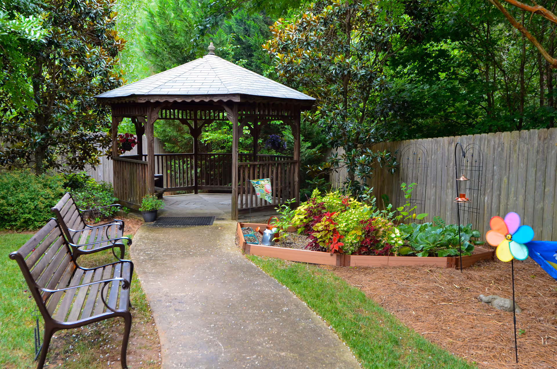 A peaceful outdoor garden area featuring a wooden gazebo with a shingled roof at the end of a paved pathway. Two wooden benches with metal armrests are positioned along the left side of the path. On the right side, there is a raised garden bed filled with colorful plants and flowers, a bird feeder, and a colorful pinwheel. The area is surrounded by green trees and a wooden fence in the background.