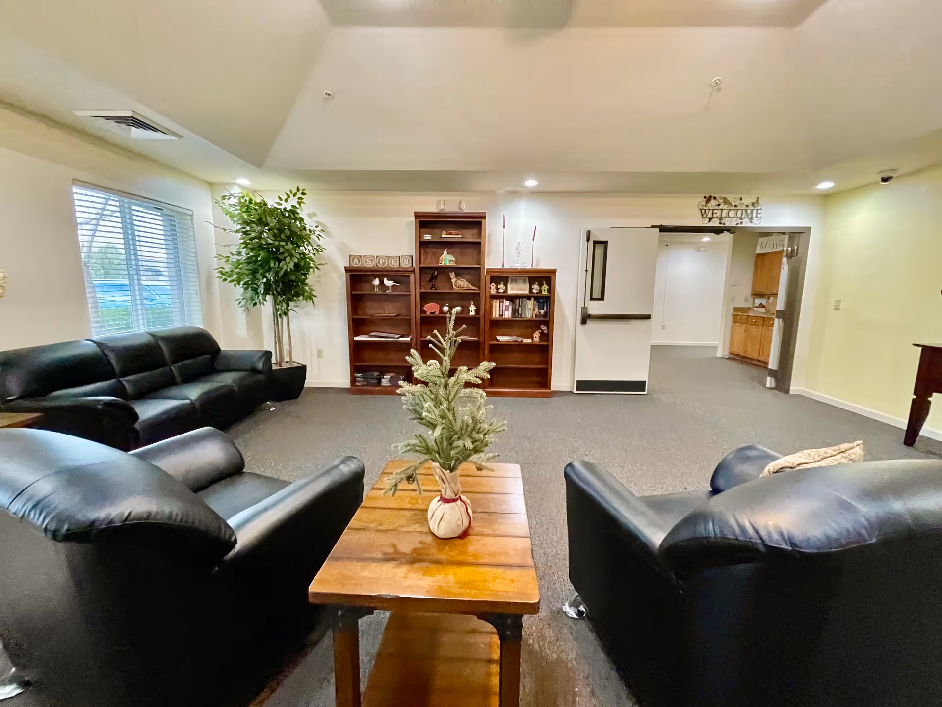 A cozy living room area with black leather sofas and armchairs arranged around a wooden coffee table with a small potted plant. Behind the seating area are three wooden bookshelves with decorative items and books. A large window with blinds is on the left wall, and a doorway with a 'Welcome' sign above it leads to another room with wooden cabinets.