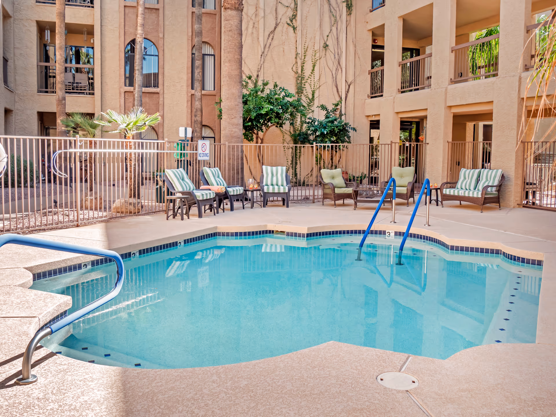 Small outdoor swimming pool in a courtyard with lounge chairs, palm trees, and surrounding apartment balconies.