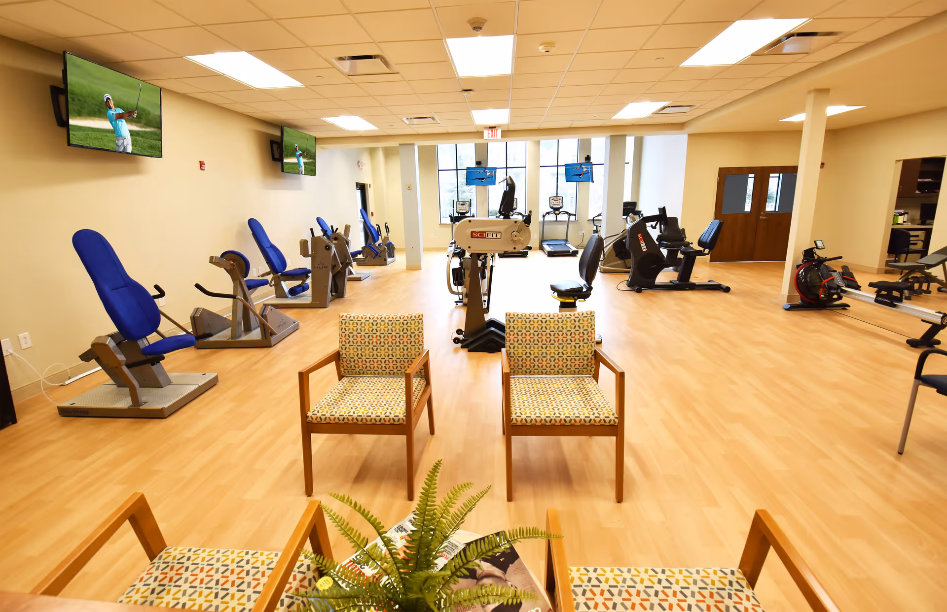 A spacious fitness room in Laurel Lake Retirement Community featuring multiple exercise machines with blue seats arranged along the left wall, several televisions mounted on the walls displaying a golf game, and a few patterned armchairs with wooden frames in the foreground. The room has light wood flooring, large windows at the far end, and bright overhead lighting.