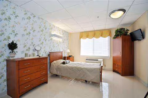 Bedroom in a senior facility with a bed, dressers, window, floral wallpaper, and wall-mounted TV.