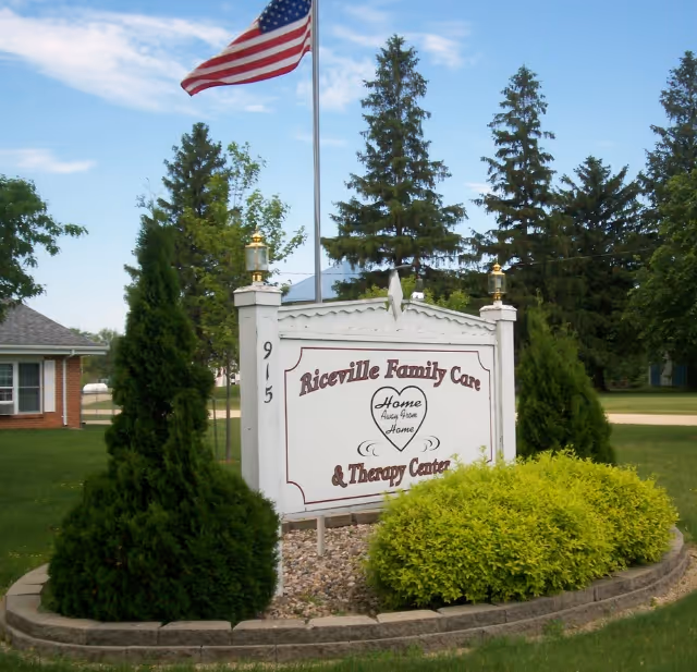 A white entrance sign for Riceville Family Care & Therapy Center surrounded by shrubs, with an American flag and tall trees in the background.