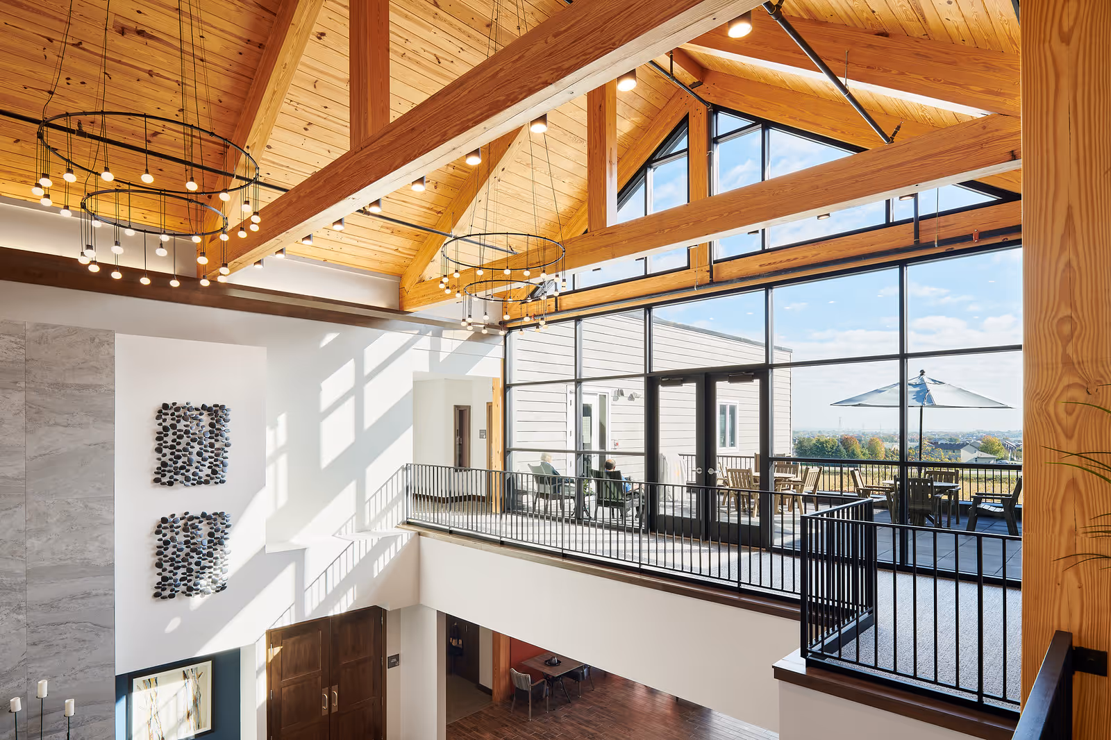 Interior view of a senior living community with a high wooden vaulted ceiling, modern circular chandeliers, large windows letting in natural light, and an upper-level balcony overlooking an outdoor patio with tables and umbrellas.