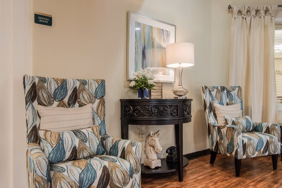 Two patterned armchairs with leaf designs flank a black ornate console table holding a lamp, a vase with white flowers, and decorative items. A framed abstract painting hangs on the wall above the table. A small sign on the wall reads 'DINING ROOM'.