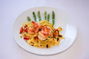 A white plate with a serving of pasta topped with shrimp and garnished with chopped vegetables, accompanied by a row of asparagus spears.