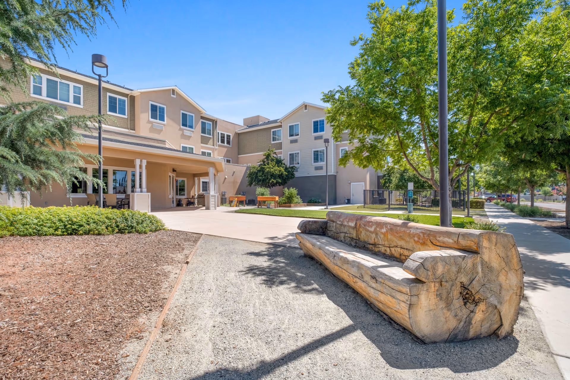 Outdoor area of Carmel Village senior living facility featuring a large wooden bench made from a single log, surrounded by trees and greenery. The multi-story building with beige and light brown exterior walls is visible in the background under a clear blue sky.