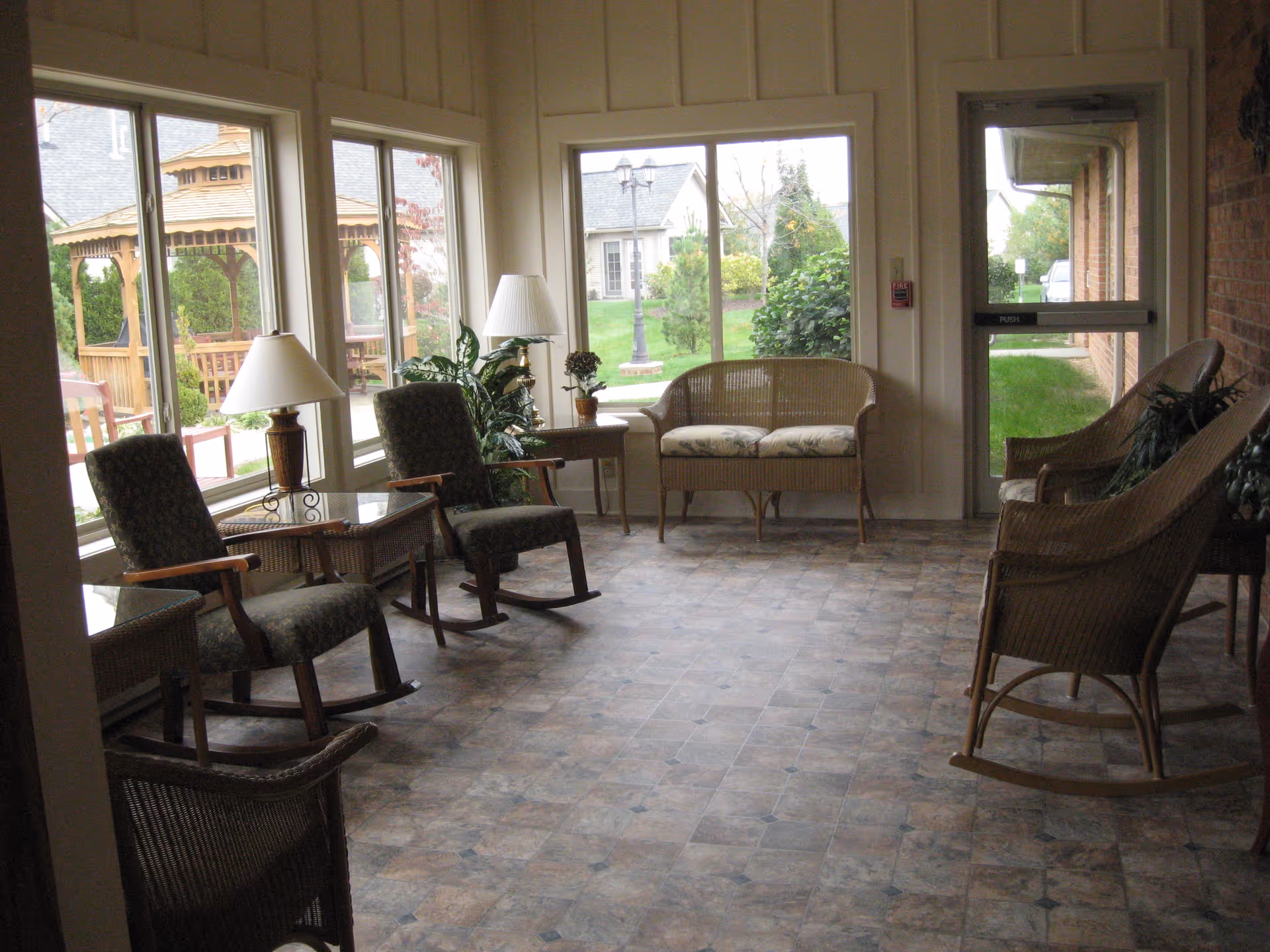 Sunlit sitting room with wicker and upholstered rocking chairs and a loveseat arranged by large windows overlooking a garden gazebo.