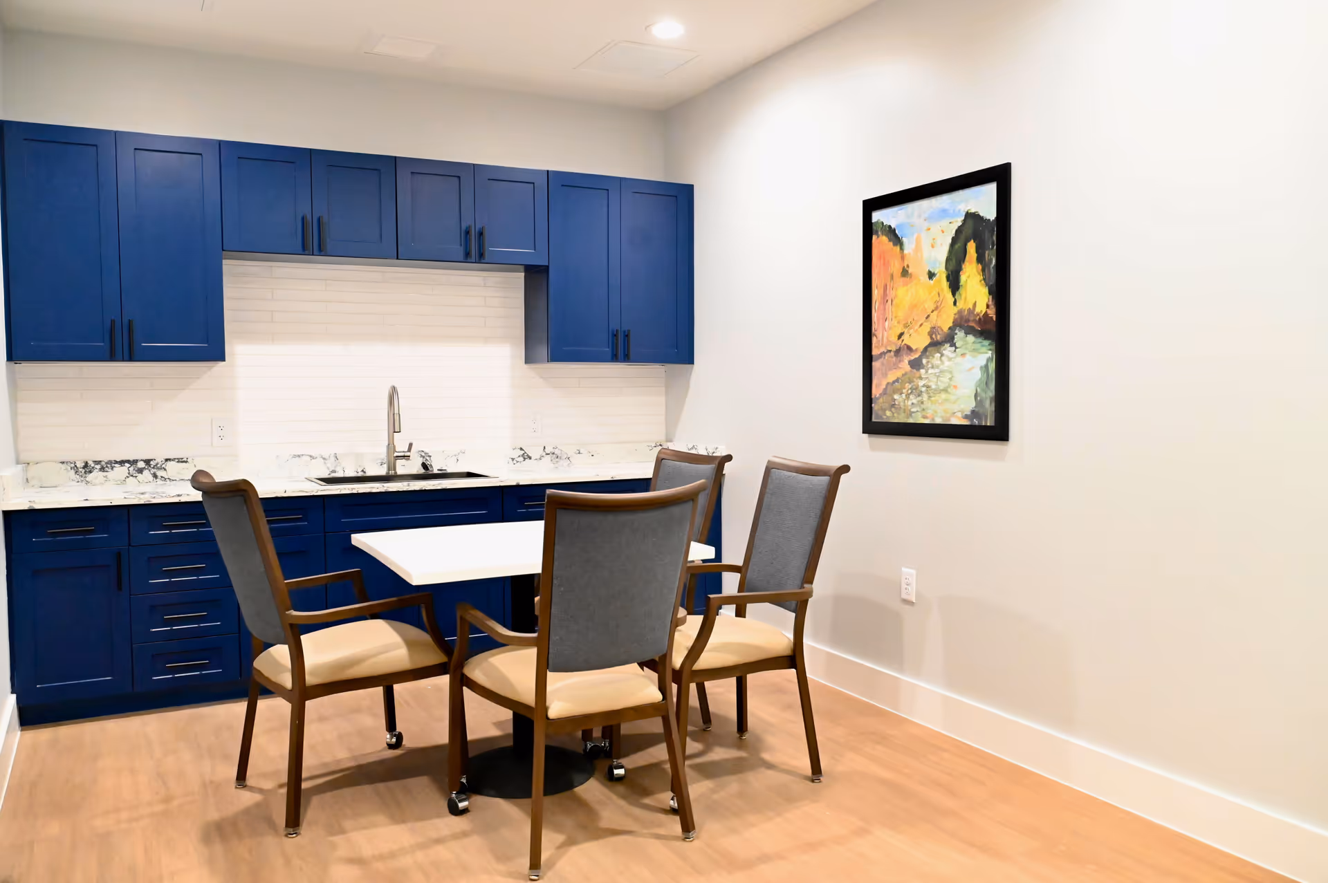 A small dining area with a white table and four chairs with wooden frames and cushioned seats. Behind the table is a kitchen area with blue cabinets, a white marble countertop, a sink, and a white tiled backsplash. A colorful framed painting hangs on the plain white wall to the right.