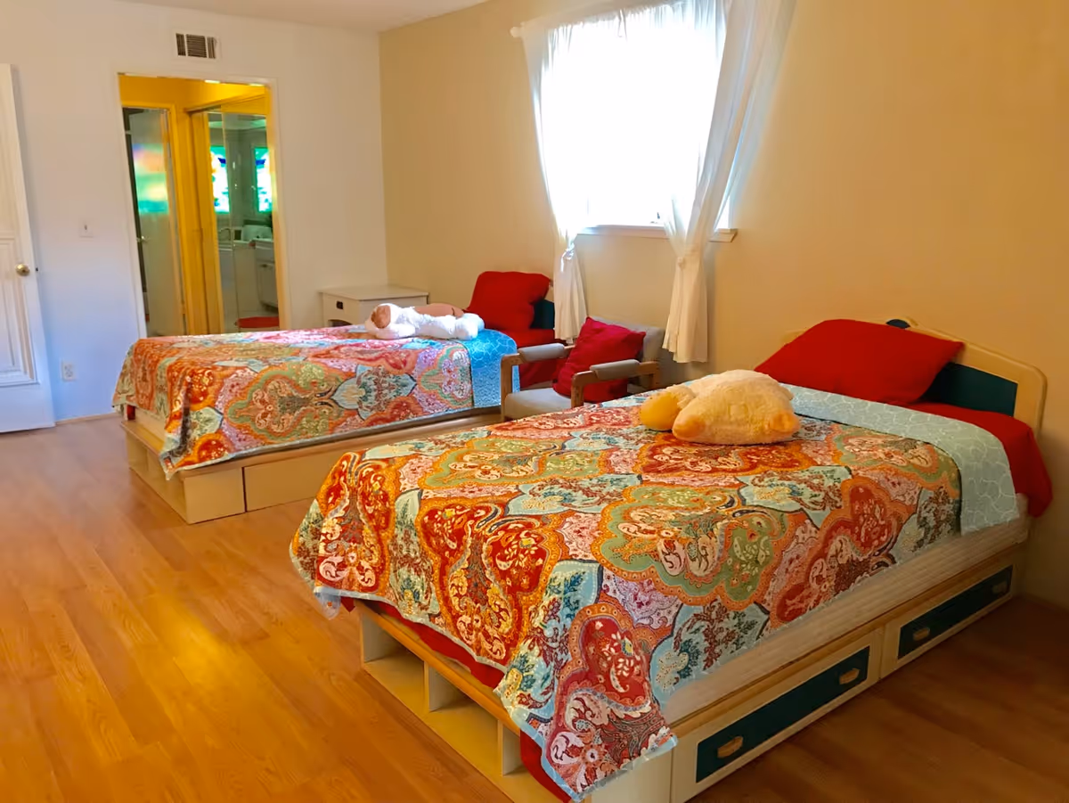 Two single beds with colorful patterned quilts and a small seating area in a bright, simply furnished bedroom.