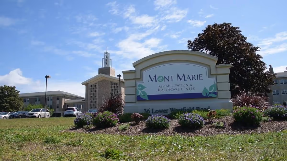 Front sign reading 'Mont Marie Rehabilitation & Healthcare Center' with the facility building and landscaped lawn behind it under a blue sky.