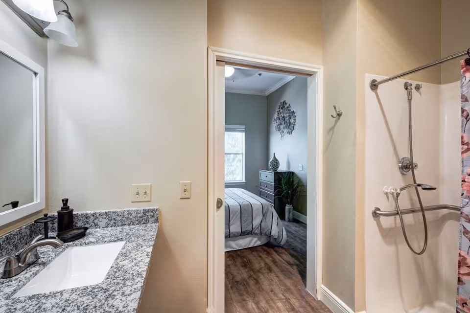View of a bathroom with a granite countertop sink and a shower with a floral curtain. Through an open doorway, a bedroom with a bed, dresser, and wall decoration is visible.