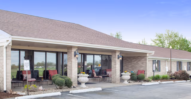 Front exterior of a single-story healthcare facility with a covered porch, outdoor chairs, planters, and adjacent parking spaces.