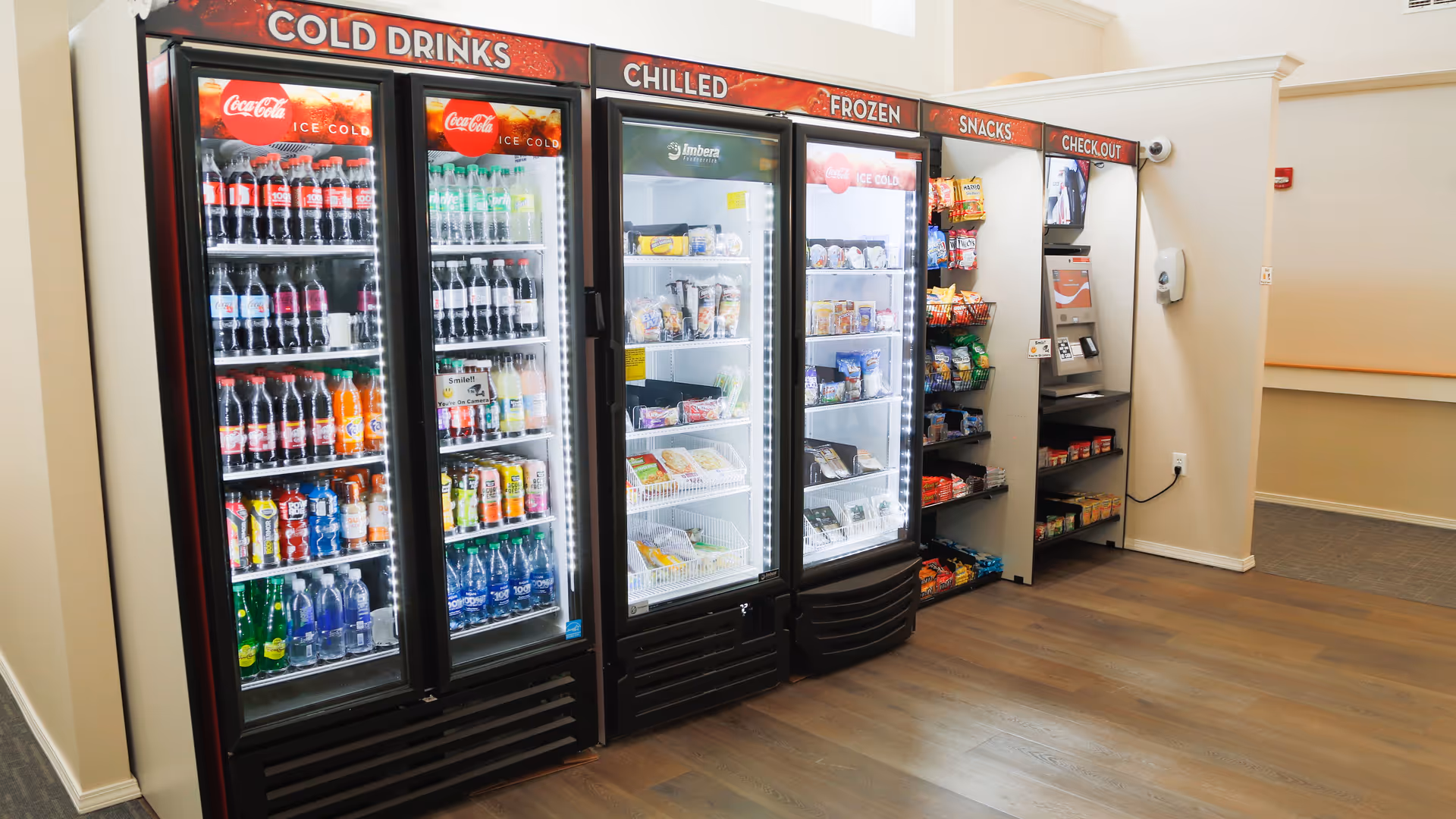 A small convenience area inside a facility featuring refrigerated glass-door coolers stocked with cold drinks, chilled and frozen food items, and shelves with snacks. There is a self-checkout kiosk on the right side and a hand sanitizer dispenser mounted on the wall.