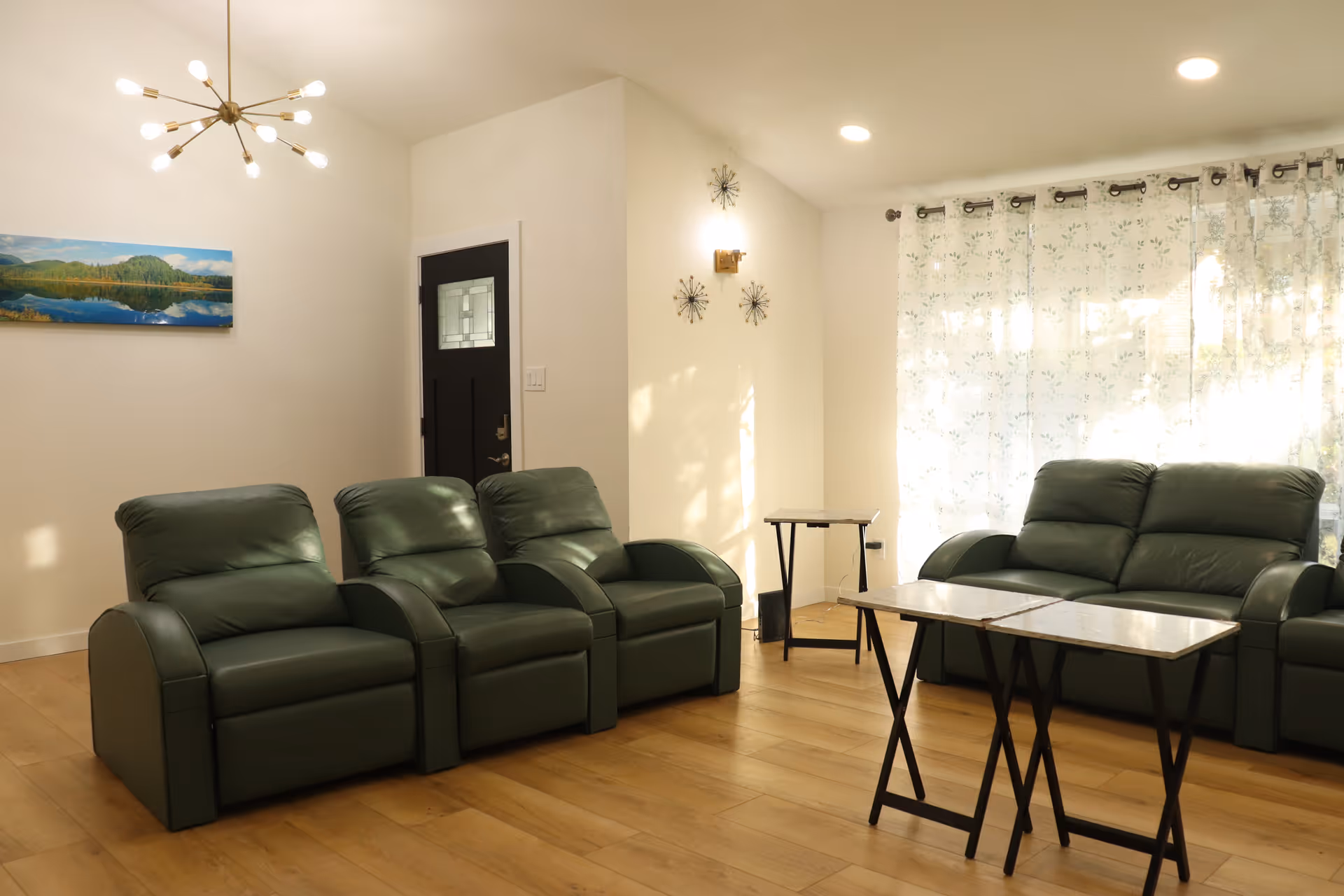 A well-lit living room with three green leather recliners and a matching green leather sofa. There are two small tables with marble tops in front of the sofa. The room has light wooden flooring, a black door with a glass panel, a modern chandelier, and sheer floral curtains covering a large window. Wall decorations include a landscape painting and three decorative metal wall pieces.