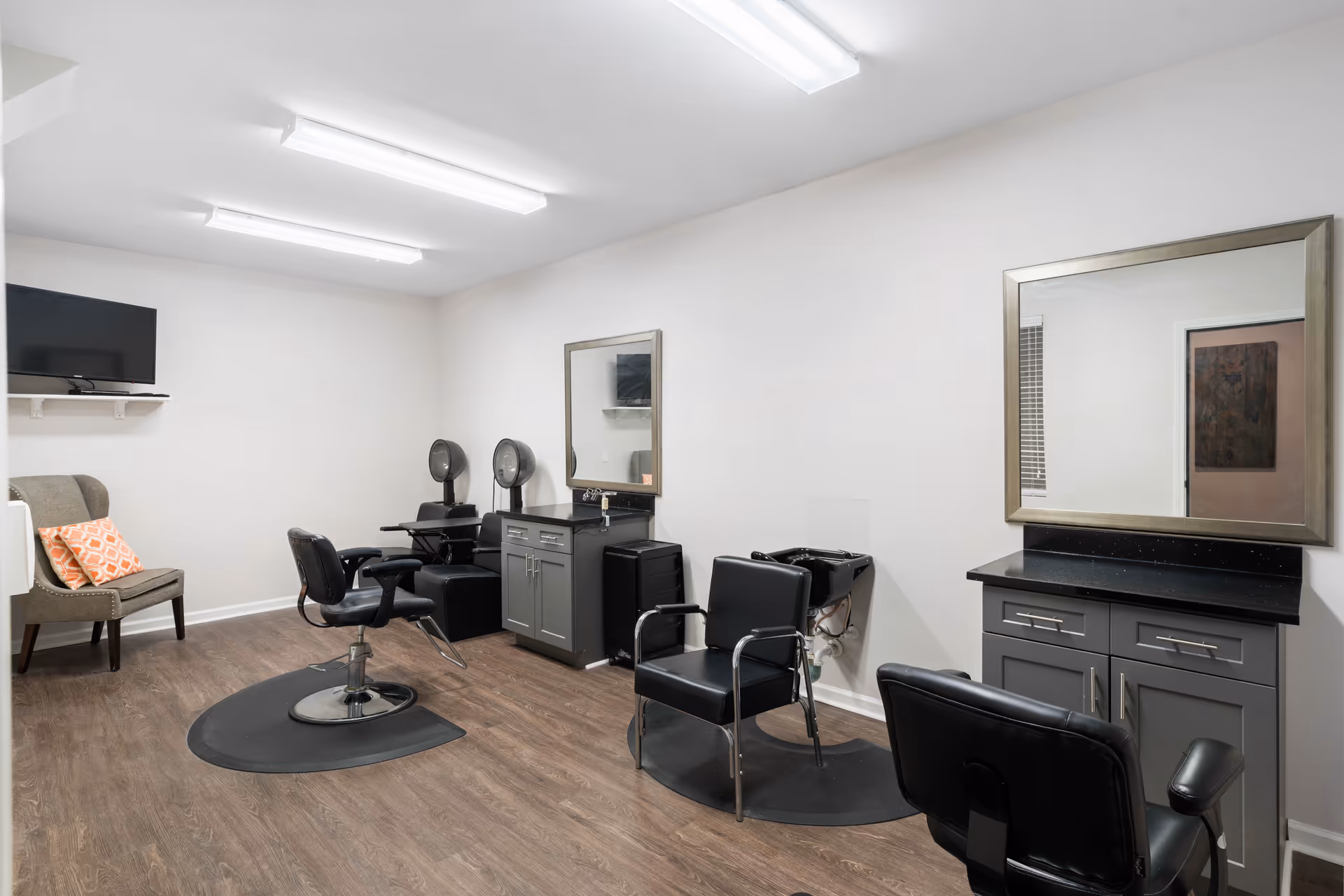 Interior view of a hair salon area in a senior living facility with salon chairs, hair drying stations, large mirrors, a small TV mounted on the wall, and a cushioned chair with an orange patterned pillow. The room has wood flooring and bright overhead lighting.